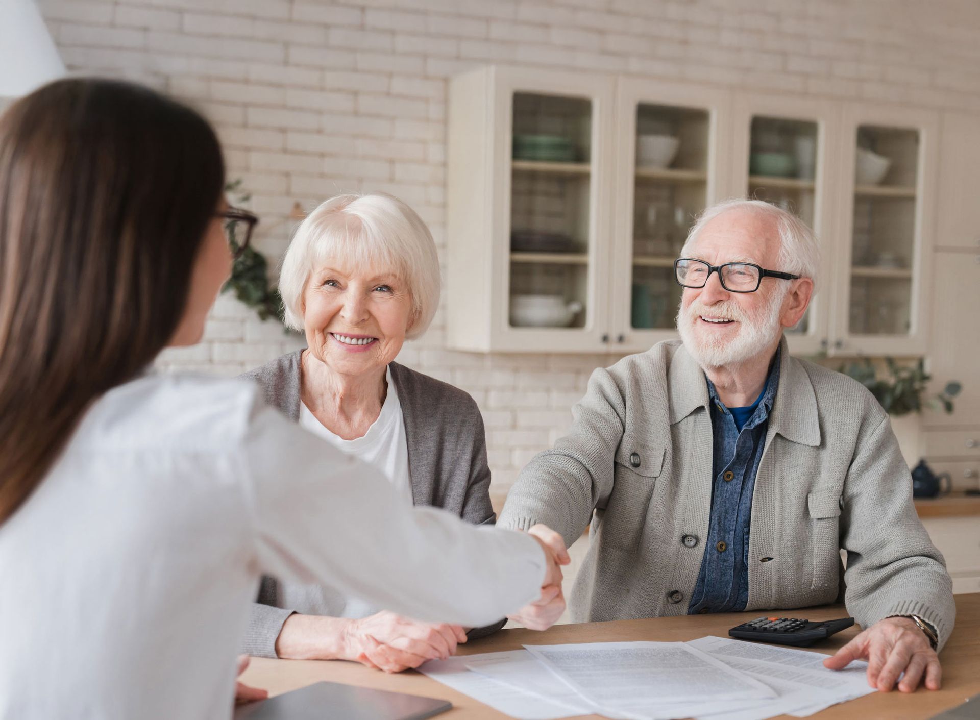Woman shaking hands with an elderly man at a table; woman and another elderly person smiling in a home kitchen.