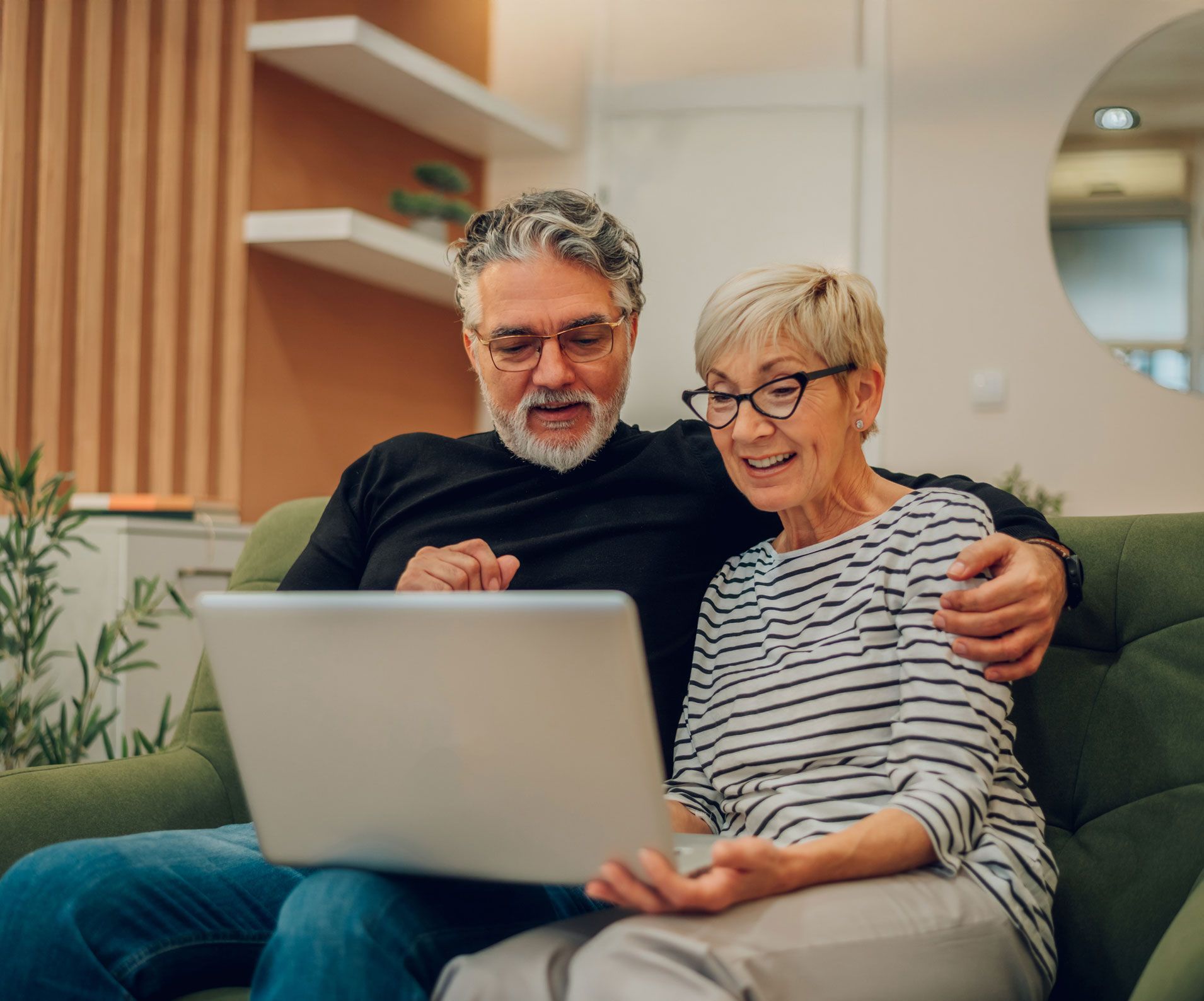 Couple on green sofa, looking at laptop screen. Man has arm around woman. Room has shelves, neutral colors.