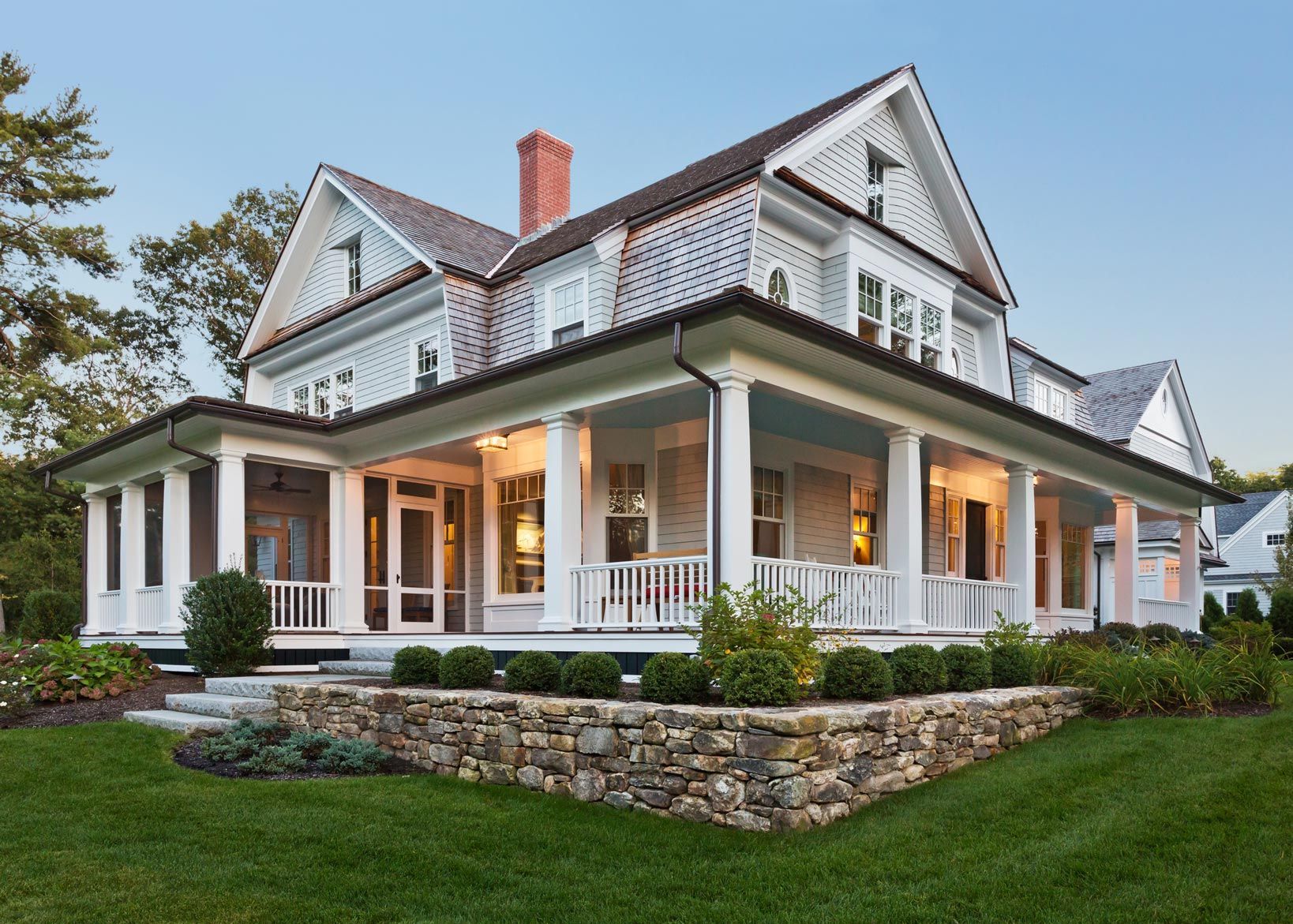 Elegant white farmhouse with wrap-around porch, stone wall, and lush green lawn.